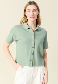 Camicia a maniche corte con colletto in morbido tessuto verde a waffle con bottoni in legno, caratterizzata da una vestibilità rilassata e dettagli di cucitura in vista.