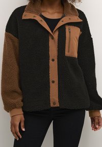 Fleecetakki, jossa mustia ja ruskeita paneleita, vuorattu kaulus, etutarrakiinnitys ja vetoketjullinen rintatasku. Pehmeä tekstuuri, jossa näkyvät saumaukset.