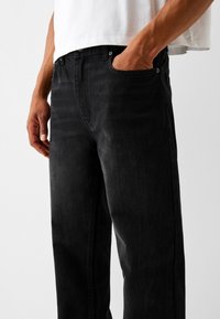 Jeans de mezclilla negra con diseño de pierna recta, estilo de cinco bolsillos y una textura ligeramente descolorida. Incluye detalles de herrajes metálicos.