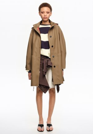 Persona con un impermeable beige abotonado sobre un suéter de rayas y chaqueta marrón con cordones, con una falda blanca de encaje y sandalias negras de punta abierta.