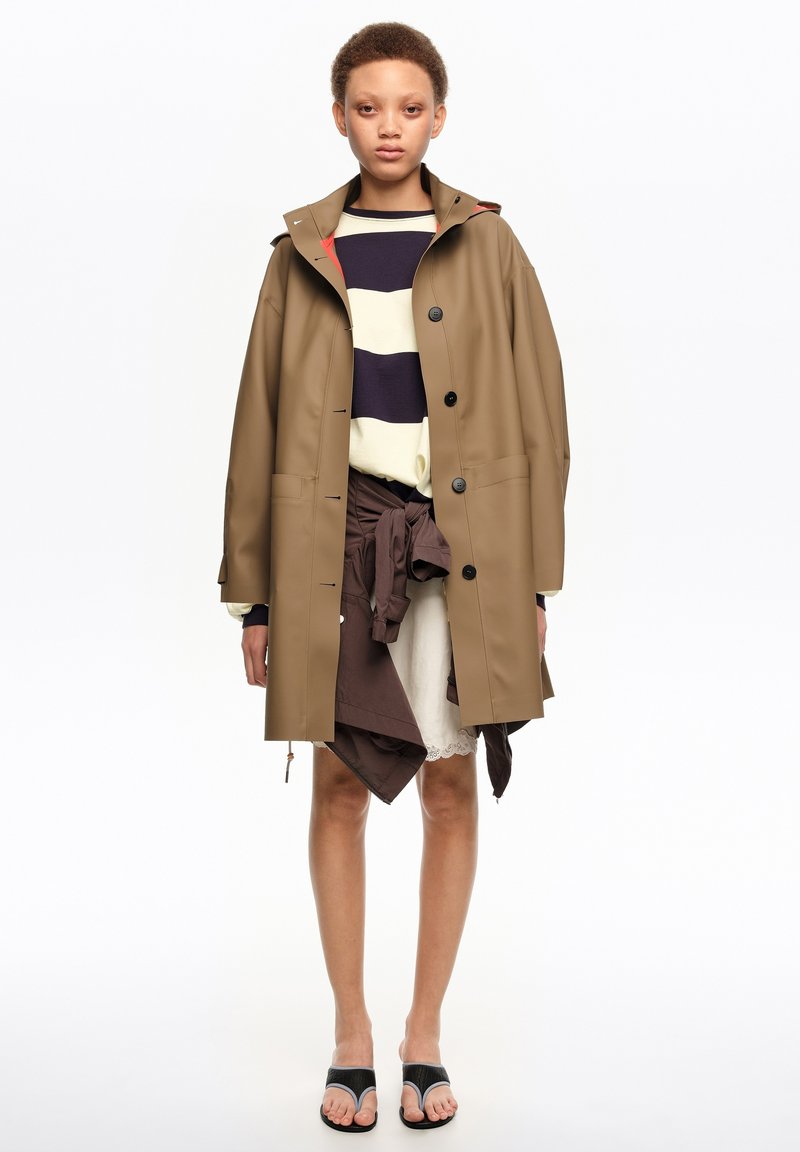 Person wearing a tan buttoned raincoat over a striped sweater and brown tied jacket, with white lace skirt and black open-toe sandals.