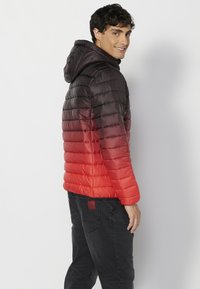 Giubbotto imbottito con un colore sfumato che passa dal nero al rosso, dotato di cappuccio e texture trapuntata, abbinato a jeans neri.
