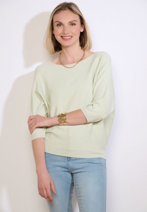 Kobieta o krótkich blond włosach, ubrana w jasny zielony sweter, niebieskie dżinsy, złoty łańcuszek i masywną złotą bransoletkę, uśmiecha się z założonymi skrzyżowanymi ramionami.