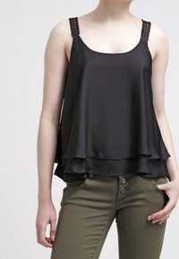 Camisole en satin noir à design superposé, bretelles perlées et coupe décontractée, associée à un pantalon vert olive avec détail de boutons.