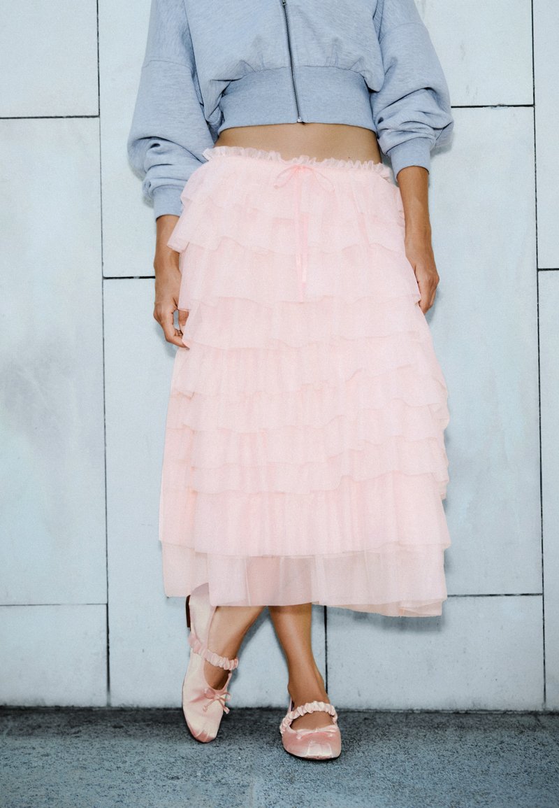 Personne portant un sweat-shirt gris court, une jupe en tulle rose superposée et des ballerines roses, debout contre un mur clair carrelé.