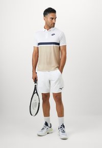 Herretøj til tennis bestående af en hvid polo med beige og marineblå vandrette striber, hvide shorts og tennissko med marineblå og grønne detaljer.