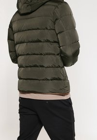 Polstret olivengrøn jakke med hætte, der har et quiltet design og en glat overflade, parret med en lysefarvet top og mørke bukser.