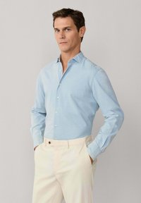 Muž stojí s rukami vo vreckách, oblečený v svetlomodrej košeli na gombíky a krémových nohaviciach na jednoduchom pozadí.