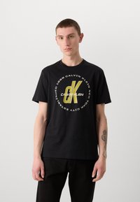 Melns kokvilnas T-krekls ar lielu dzeltenu un baltu apaļu logo, kurā ir uzraksts "Calvin Klein", ar īsām piedurknēm un apaļu kakla izgriezumu.
