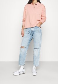 Hellrosa langärmliges Polo mit Kragen, Tasche und drei Knöpfen. Kombiniert mit hellblauen Jeans mit Used-Effekt und weißen High-Top-Sneakern.