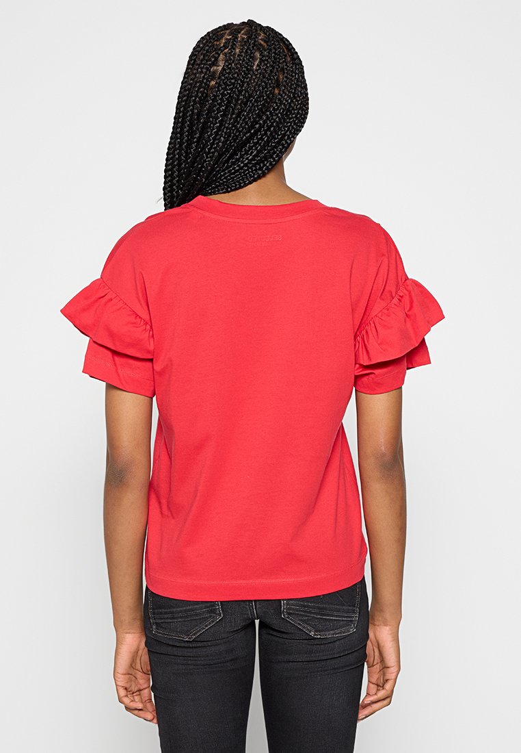 Rotes Baumwoll-T-Shirt mit kurzen, Rüschensleeves und einem runden Ausschnitt. Die Rückseite ist schlicht, kombiniert mit schwarzen, schmalen Jeans.