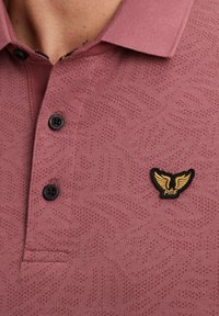 Polo à manches courtes en rose poussiéreux, avec une texture, comportant une braguette à deux boutons et un patch logo noir avec des accents dorés.