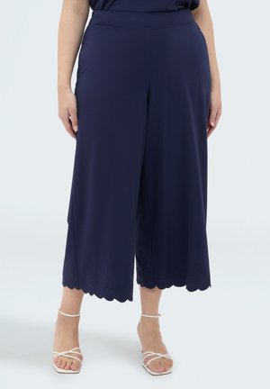 Kobieta ubrana w granatowe szerokie, krótkie spodnie z falistym brzegiem oraz białe sandały na obcasie ze paskami, stojąca na jednolitym tle.