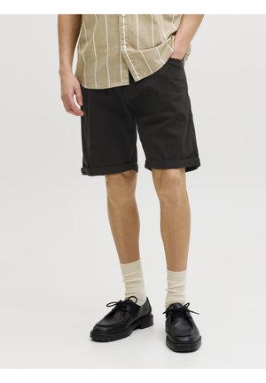 Homme portant une chemise beige rayée, un short noir retroussé, des chaussettes écrues et des chaussures à lacets noires, debout devant un fond blanc.