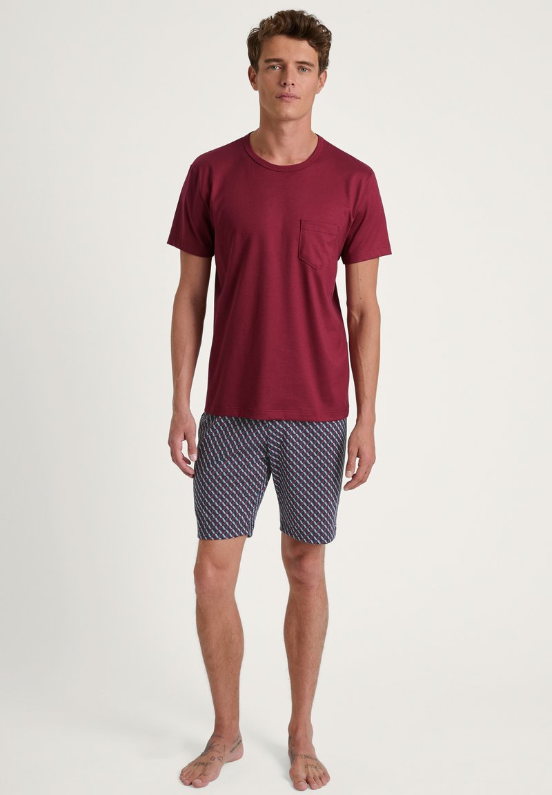 Weinrotes T-Shirt mit kurzen Ärmeln und Brusttasche, kombiniert mit gemusterten grau-blauen Shorts, die rote und blaue geometrische Designs aufweisen.