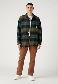 Grünes und blaues kariertes Flanellhemd mit durchgehender Knopfleiste und Brusttasche, kombiniert mit braunen Cargo-Hosen und weißen Sneakers.