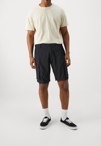 Shorts cargo noirs avec poches latérales, fermeture à bouton et coupe décontractée, associés à un t-shirt beige clair et des baskets noires.