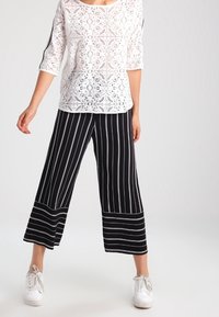Top in pizzo bianco con maniche a tre quarti, abbinato a pantaloni a gamba larga a righe bianche e nere, completato da sneakers bianche.