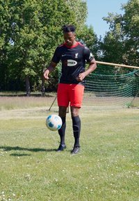 Camiseta de fútbol negra con acentos rojos, pantalones cortos rojos, calcetines negros. El jugador controla un balón de fútbol azul y blanco en un campo de hierba con una portería al fondo.