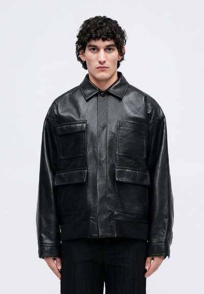 Homme portant une veste en cuir noir avec de grandes poches frontales, se tenant devant un fond gris clair uni, faisant face à l'avant.