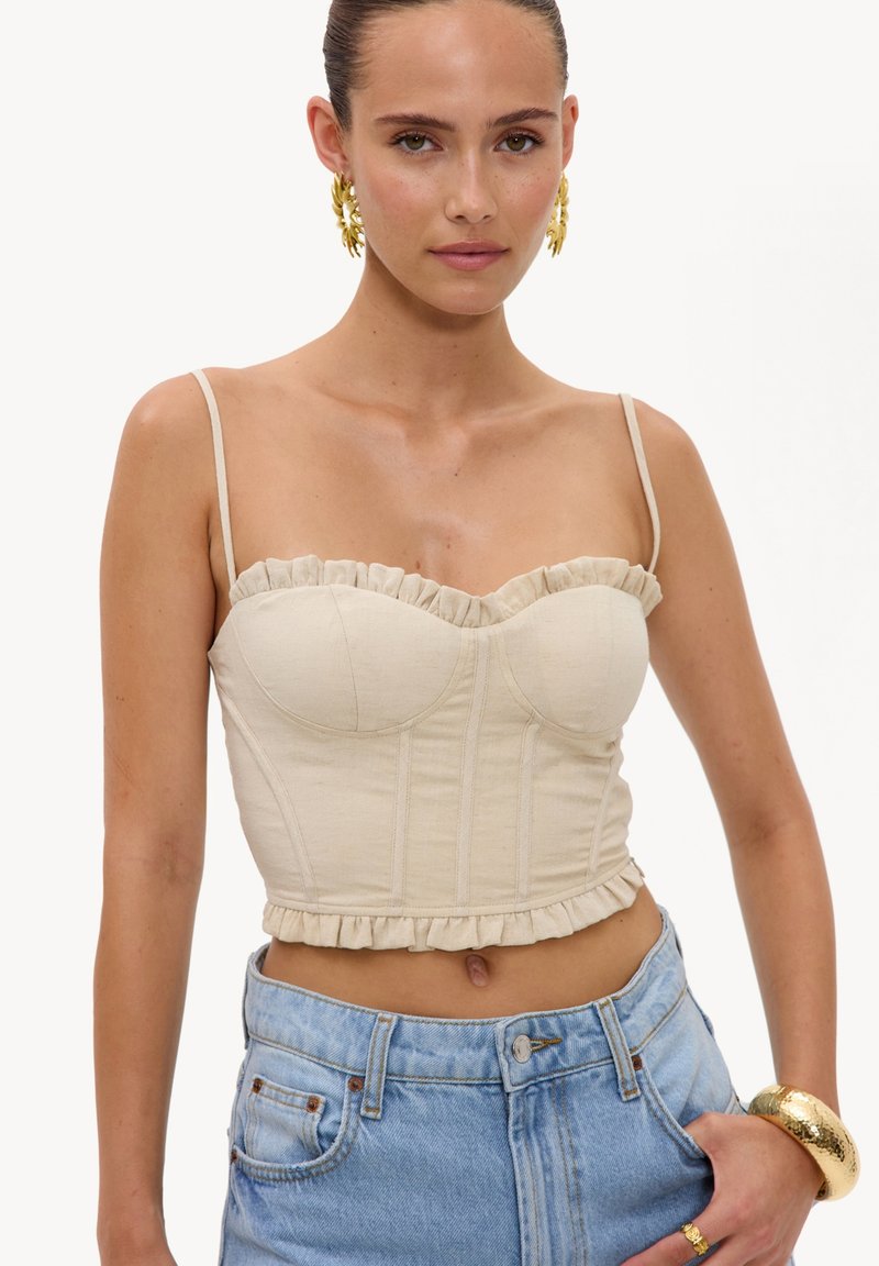 Femme portant un crop top beige à volants avec fines bretelles, un jean taille haute bleu clair, des boucles d’oreilles créoles dorées, une bague dorée et un bracelet doré.