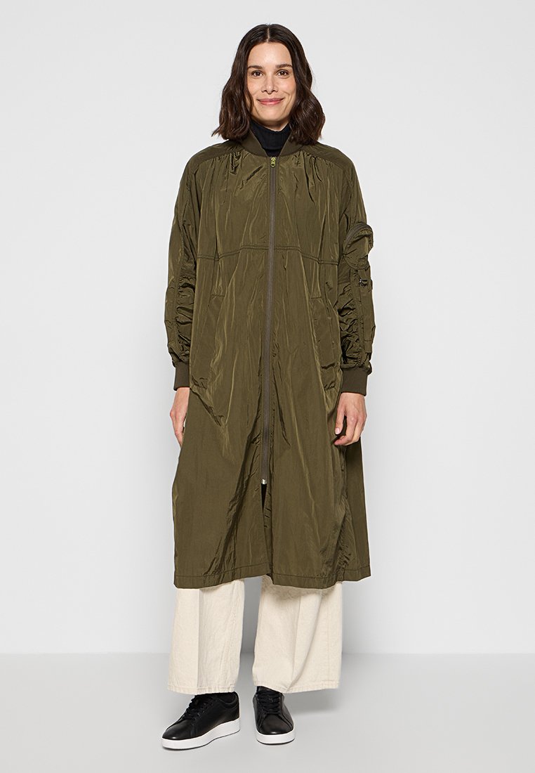 Oliwkowa długa kurtka z zamkiem błyskawicznym, z marszczonymi rękawami i bocznymi kieszeniami, wykonana z lekkiej, teksturowanej tkaniny. Noszona na kremowe spodnie wide-leg.