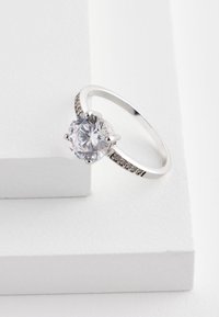 Silberner Ring mit einem großen, runden, klaren Edelstein in einer vierzackigen Fassung und kleineren Steinen, die am Ringband eingelassen sind.