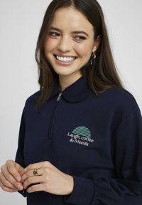 Sweat-shirt bleu marine avec un col zippé, présentant un graphique circulaire à rayures vertes et le texte "Rires, café et amis", ainsi que des manches froncées.