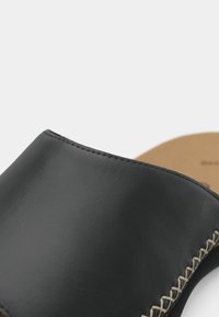 Chaussure noire en cuir à enfiler avec semelle intérieure beige et surpiqûres beiges le long de la couture latérale, présentée en gros plan.