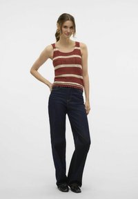 Rood en crèmekleurig gestreept mouwloos shirt met gekartelde zoom, gecombineerd met donkerblauwe wijde jeans. Model staat met één hand op de heup.