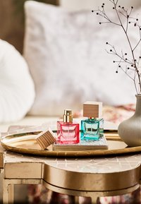 Deux flacons de parfum en verre carré avec des accents dorés, un rose et un bleu, sur un plateau décoratif avec un couvercle en bois et un carrelage texturé en dessous.
