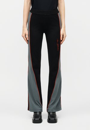 Personne porte un pantalon évasé noir et gris avec un passepoil rouge et un script rouge sur la cuisse, associé à des chaussures noires, debout sur un fond blanc.