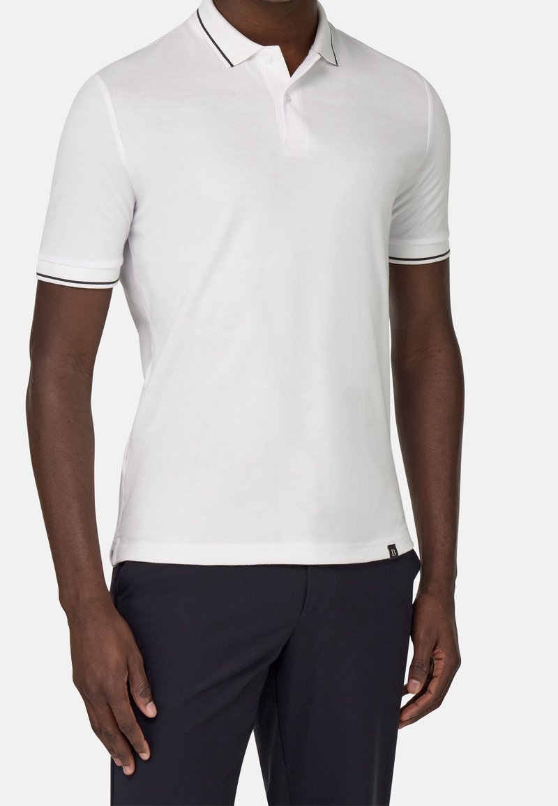Homme portant un polo blanc à manches courtes avec un liseré noir au col et aux poignets, associé à un pantalon noir sur un fond uni.