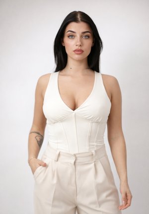 Jonge vrouw met lang donker haar, gekleed in een witte mouwloze top en beige hoge taille broek, staand tegen een effen lichte achtergrond.