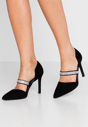 Escarpins en daim noir à talons hauts avec bouts pointus et bride décorée de strass portés pieds nus sur un fond gris uni.