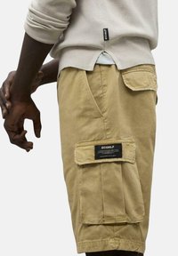 Pantalones cortos cargo color caqui con múltiples bolsillos, una etiqueta de tela y un ajuste relajado. Fabricados con material duradero, con costuras visibles y textura.