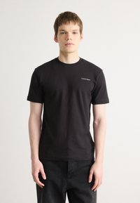 Fekete pamut póló rövid ujjakkal, kerek nyakkivágással, és kicsi fehér "Calvin Klein" logóval a bal mellrészen. A modellen sötét farmernadrág van.