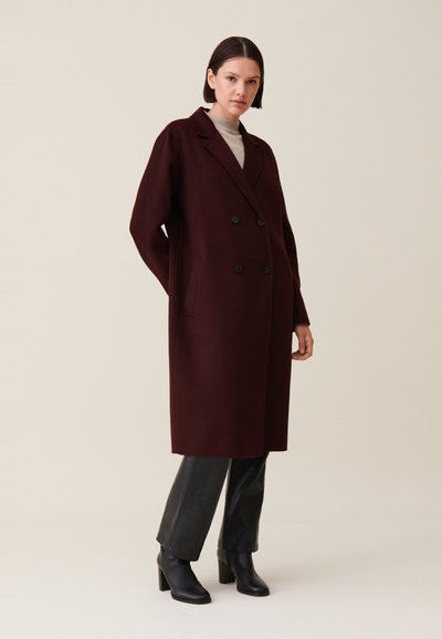 Cappotto doppiopetto bordeaux in lana, con bottoni neri e tasche laterali, abbinato a pantaloni neri aderenti e stivaletti.