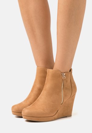 Botas de tornozelo em camurça bege com salto em cunha, apresentando um bico arredondado e detalhe de fecho lateral, com uma textura suave e um salto robusto.