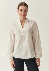 Femme aux cheveux tirés en arrière, portant un chemisier blanc à manches longues brodé de fleurs et un jean foncé sur un fond uni.