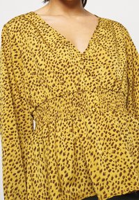 Gul blus med bruna leopardprickar, v-ringning, knappar fram, och samlad midja, med långa ärmar och mjuk tygstruktur.