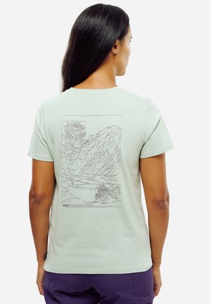 Femme aux longs cheveux bruns portant un t-shirt vert clair avec un dessin en lignes de montagnes et de fleurs dans le dos, associé à un pantalon violet.