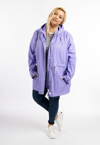 Šviesiai violetinė vandeniui atspari striukė su kapišonu, užtrauktuku, reguliuojamomis raištelėmis ir priekinėmis kišenėmis, derinama su tamsiai mėlynomis džinsomis ir baltais sportbačiais.