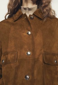 Giacca in suede marrone con bottoni a scatto argentati e due tasche a ribalta, indossata sopra un top beige con zip.