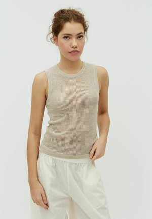 Junge Frau mit lockigem Haar trägt beiges ärmelloses Netzoberteil und weite weiße Hose, steht vor hellem einfarbigem Hintergrund.