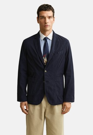 Man draagt een marineblauwe blazer, blauw gestreept overhemd, marineblauwe stropdas met zeilbootmotief en beige broek, staand tegen een effen witte achtergrond.