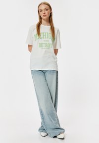 Hvid bomulds t-shirt med teksten "MICHIGAN EST. 1990 DETROIT" i grøn skrift, parret med lyseblå viddejeans og hvide sko.