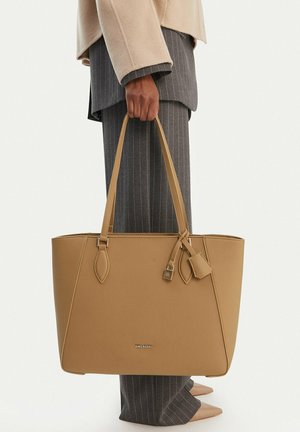 Persona con traje de raya diplomática gris y abrigo beige sosteniendo una gran bolsa tote de cuero marrón claro con detalle de cierre a un lado.