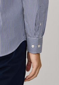 De arm en hand van een man die een blauw-witte verticaal gestreepte overhemd met knopen op de manchetten draagt, naast donkerblauwe broek.