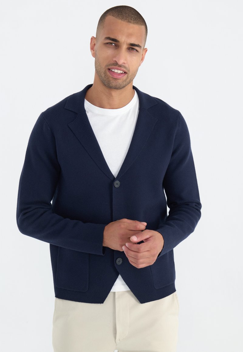 Navyblauer Strickcardigan mit Schalkragen, einer einzelnen Knopfschließe und zwei Fronttaschen, getragen über einem weißen T-Shirt. Glatte Textur.